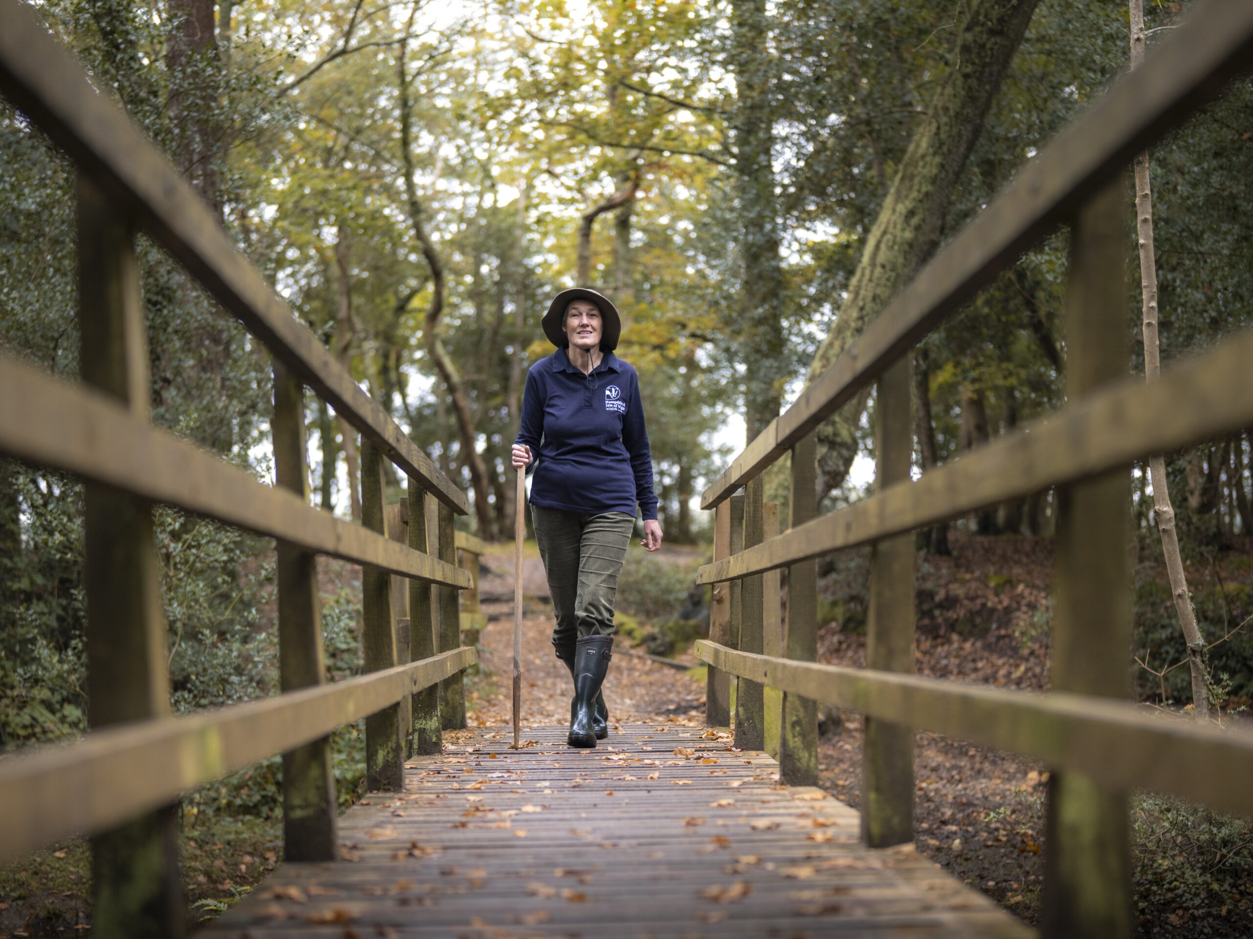 Catherine Chatters walking across a wooden footbridge in a forest, wearing a hat and holding a walking stick