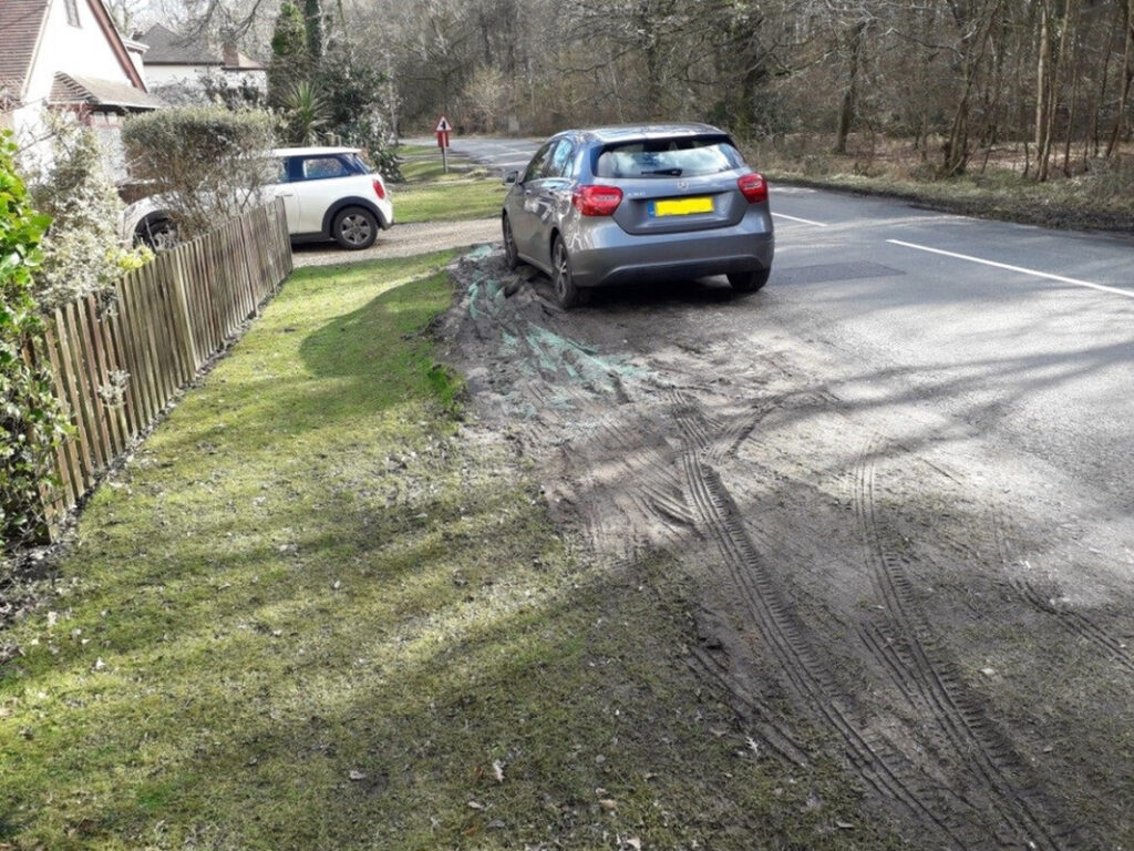Car parked partly on muddy, eroded roadside verge with tire ruts beside a paved road near houses and trees
