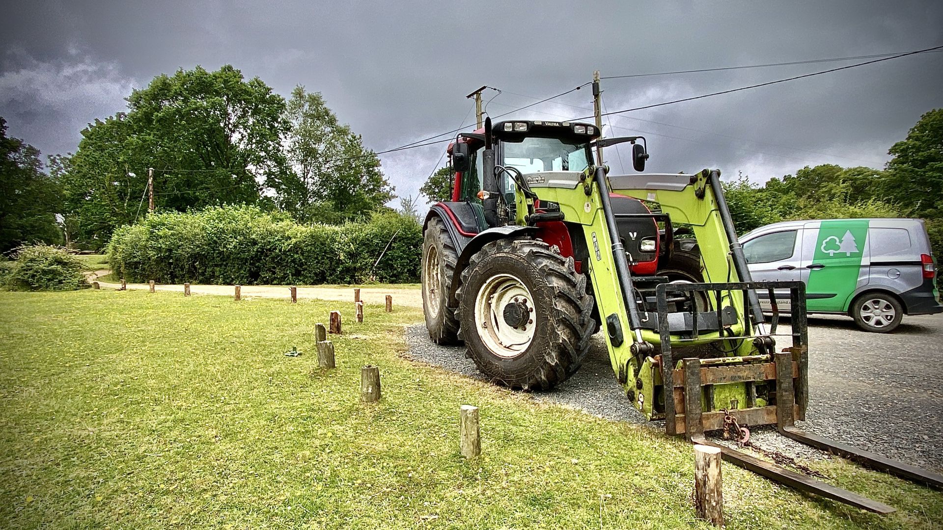 Tractor with front loader parked on gravel verge beside wooden posts, with a green-and-white van and trees under grey sky