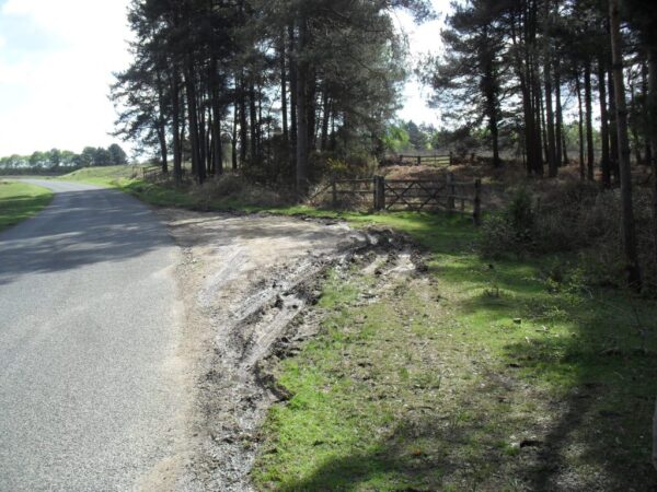 Eroded road verge with deep ruts and mud beside a paved lane, next to a wooded area and fence gate