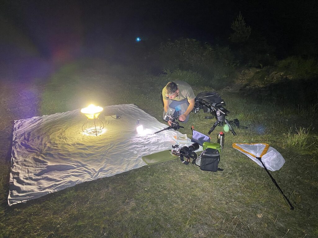Man kneels on grass at night adjusting a camera, with a lit lantern on a tarp and filming gear and tripod nearby