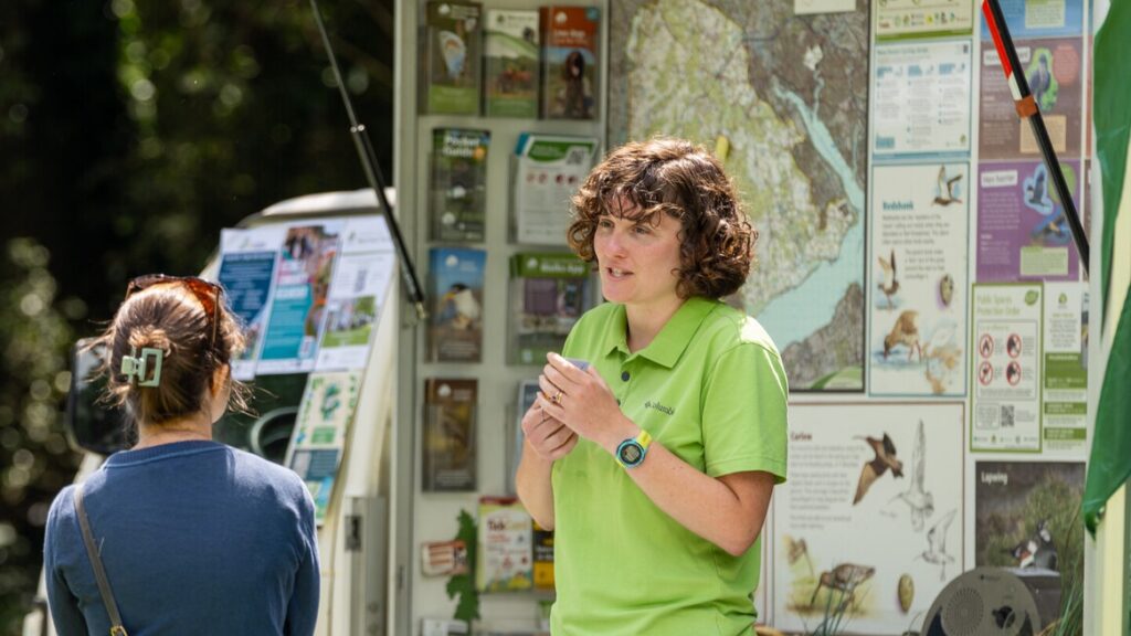 Park ranger talks with visitors at an outdoor information stand, with maps and wildlife posters on display